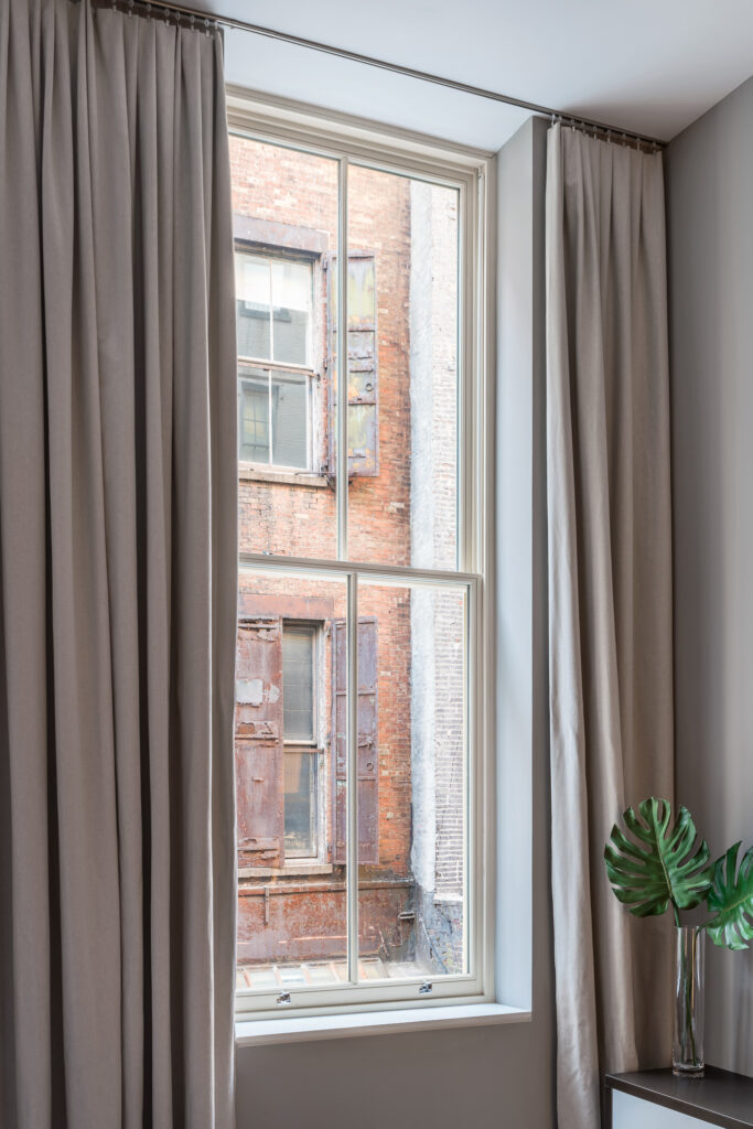 A tall, white-framed double-hung window, flanked by floor-to-ceiling pleated grey curtains, reveals a view of a weathered red-brick industrial building featuring narrow windows and rusted metal shutters, juxtaposing a clean, modern interior—complete with a vibrant green monstera leaf in a glass vase—against the textured, urban grit of the city outside.