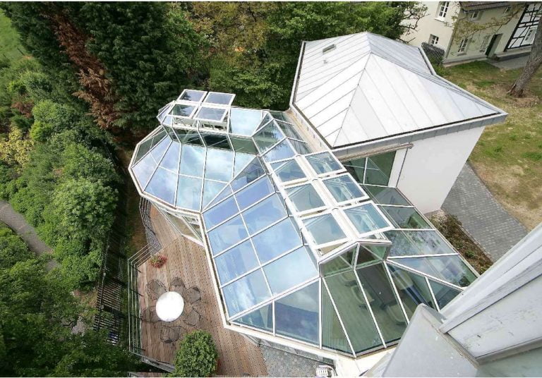 Aerial view of a modern building extension with multiple glass skylights and an angular, geometric roof design thet allows natural light to illuminate the interior, with various sections creating a greenhouse-like effect.