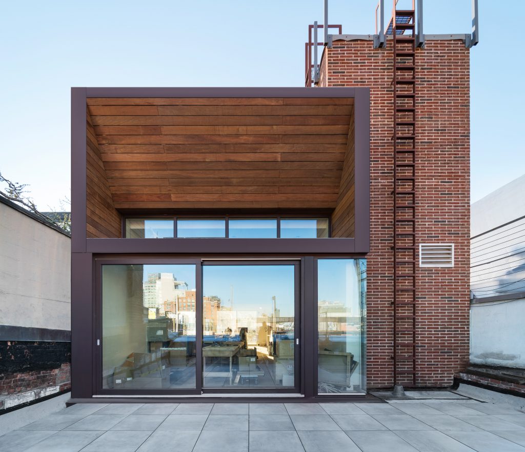 Modern rooftop structure with large glass sliding doors and wooden ceiling panels, adjacent to a brick chimney with a metal ladder, overlooking an urban skyline.