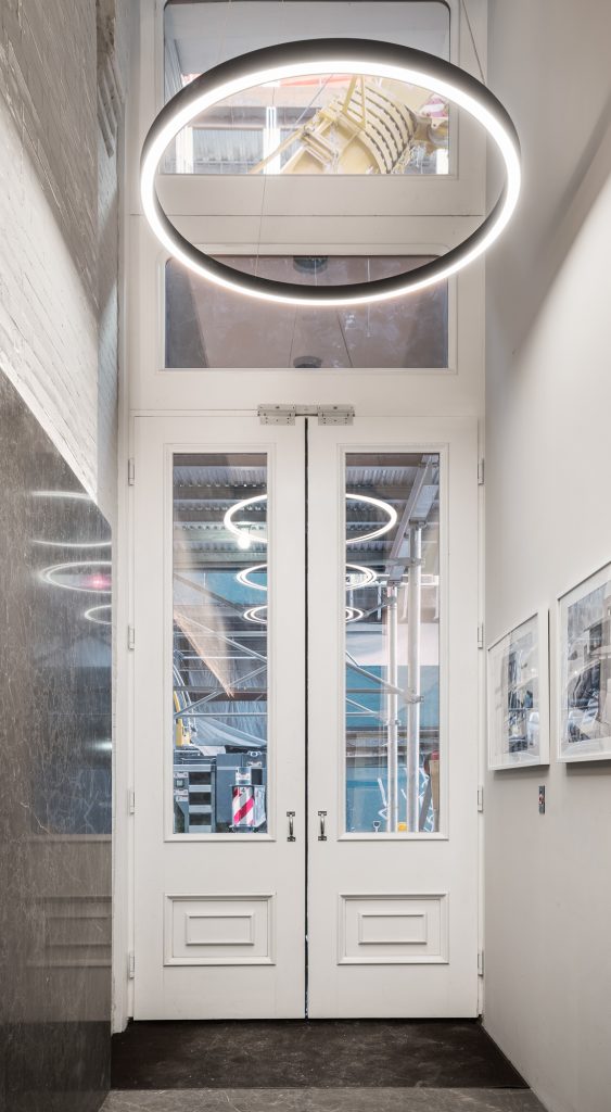 Modern double French doors with glass panels and a fixed transom, illuminated by a circular ceiling light, set in a contemporary hallway with white walls and marble accents.