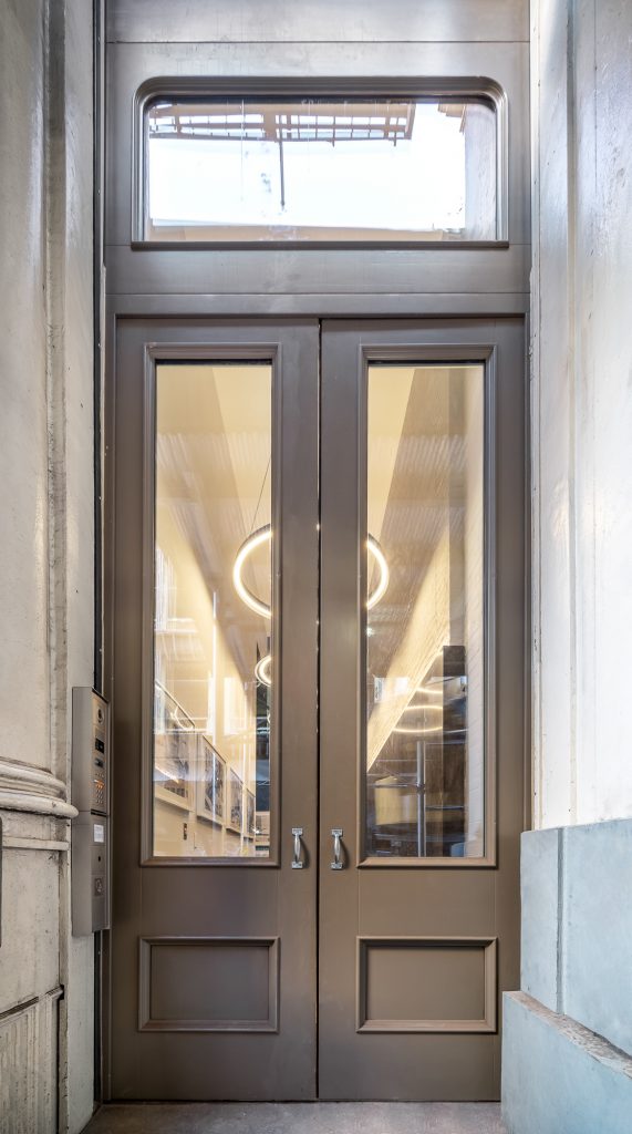 Elegant brown double French doors with glass panels and a fixed transom, leading to a brightly lit interior with visible hanging lights, in a classic architectural setting.