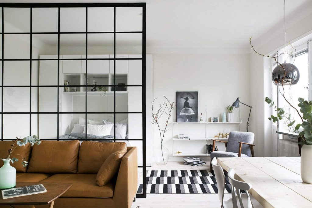 Modern living room with a black steel-framed glass partition separating the bedroom area, featuring a cozy brown sofa and minimalist decor.