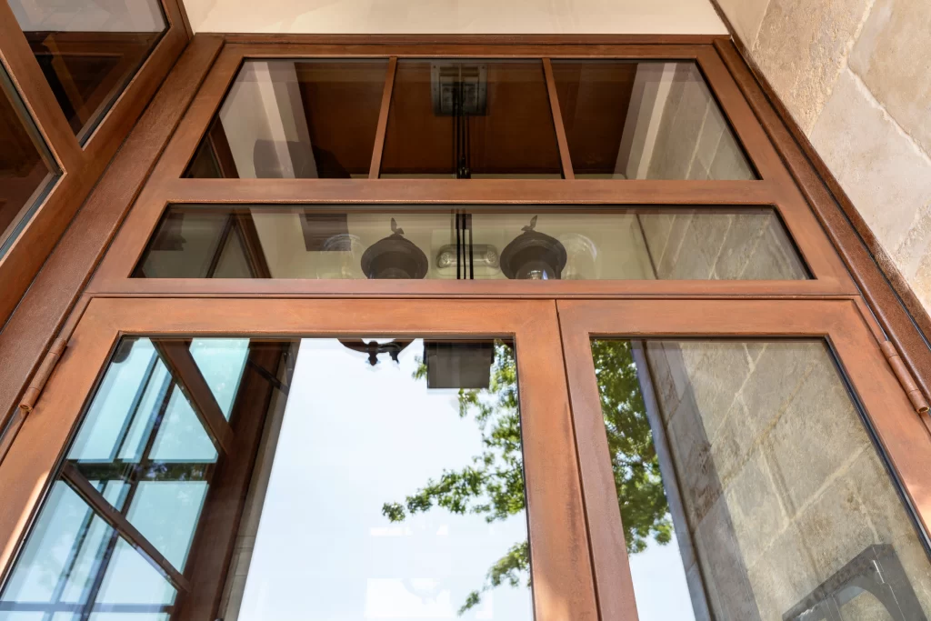 Tall, elegant window with a warm, corten steel, featuring clean and precise edges, the reflective glass panels capture a clear view of the outdoor greenery and interior lighting fixtures above, complementing the textured stone walls surrounding it.