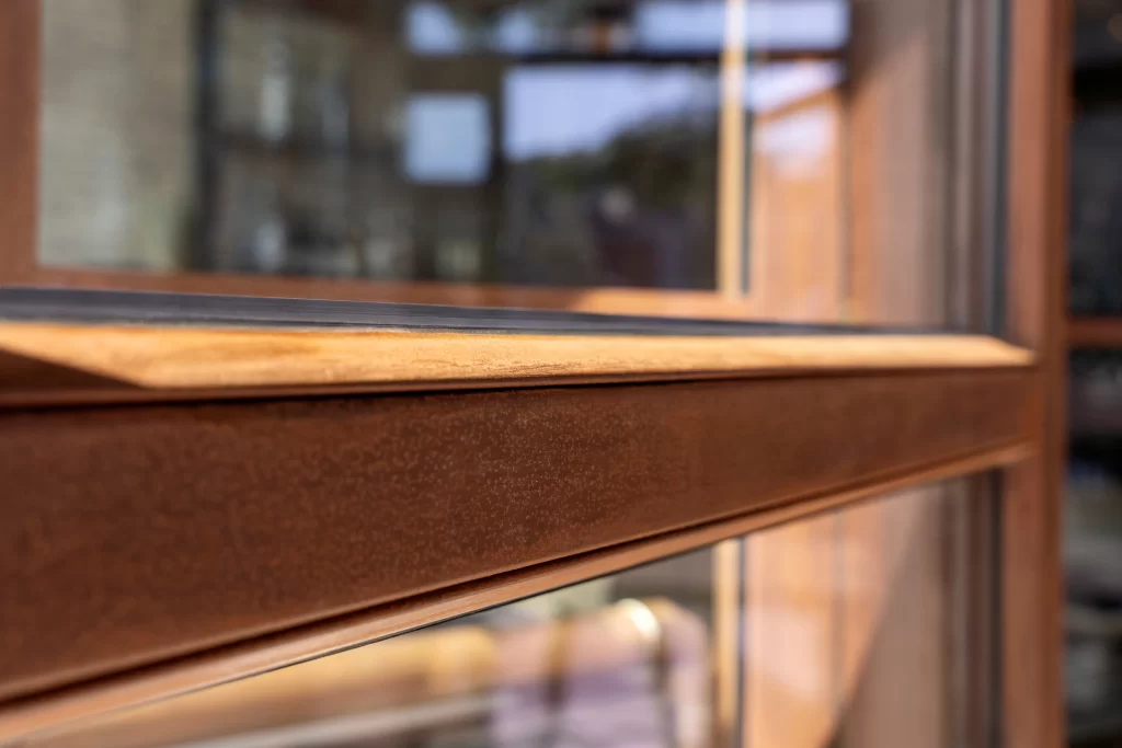 Close-up of a window frame with a warm, corten steel finish, highlighting its finely textured surface and clean edges, with the reflective glass panel offering a view of the surrounding interior.