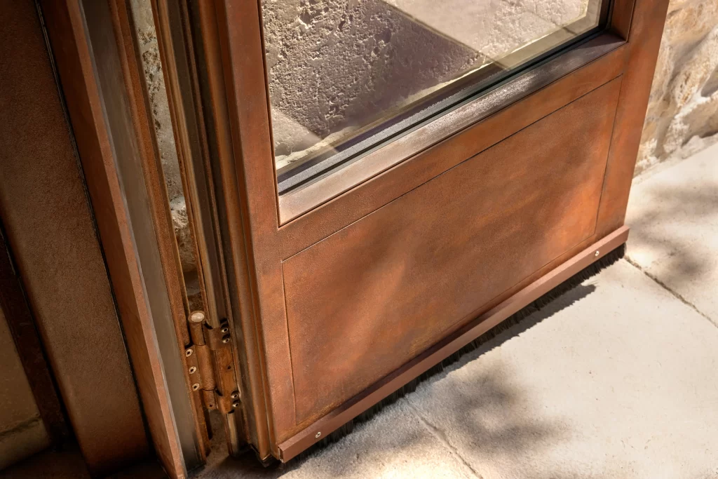 The lower section of a finely designed door with a warm, rust-toned corten steel, showcasing detailed craftsmanship around the frame, precise glass paneling, and integrated elements like a brush weather seal at the base for functionality and aesthetic balance.