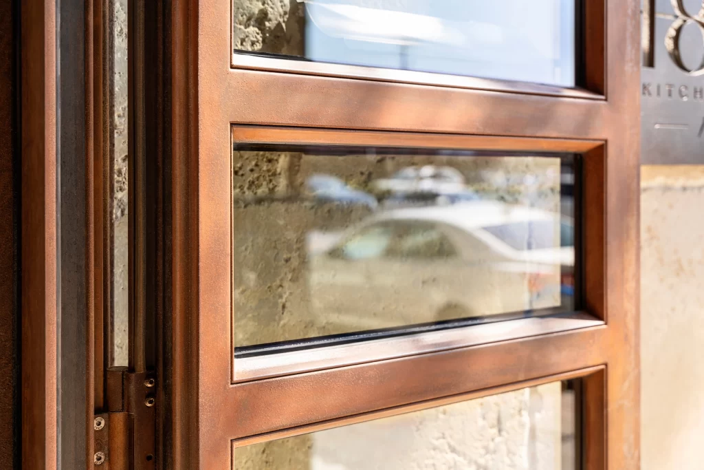 Close-up of a beautifully crafted corten steel frame with a warm, copper-like patina finish, elegantly enclosing clear glass panes, reflecting subtle outdoor details and emphasizing the refined craftsmanship and rich texture of the material.