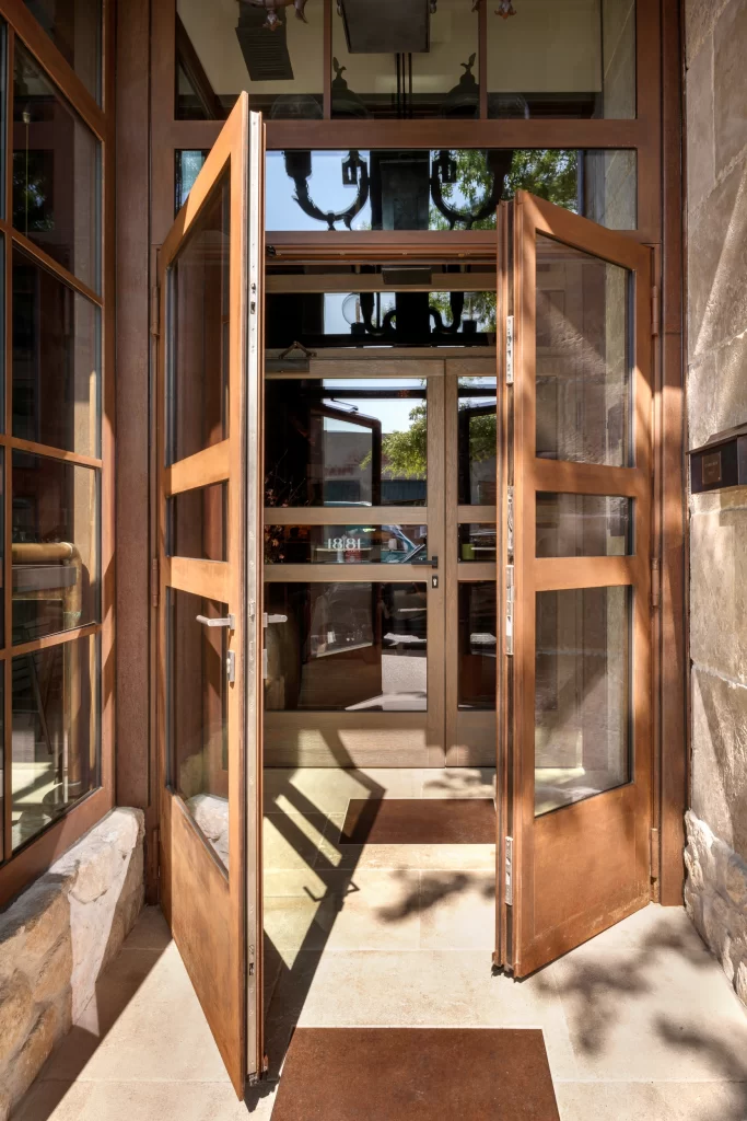 Sophisticated double-door entrance made of rich, corten steel with expansive glass panels, set against a backdrop of stone walls and bathed in sunlight, creating an inviting and elegant entryway that seamlessly connects the indoor and outdoor spaces.