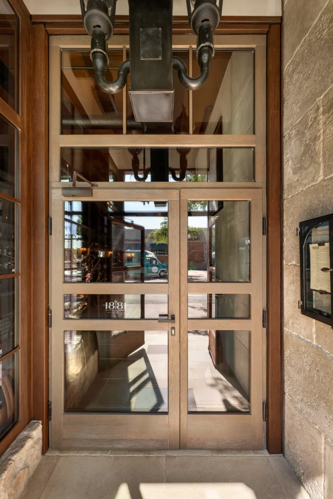 Beautifully designed entrance door with a sturdy wooden frame and large glass panels, flanked by natural stone walls; the intricate details include a modern handle and reflective surfaces that showcase the interior ambiance and the exterior street view.