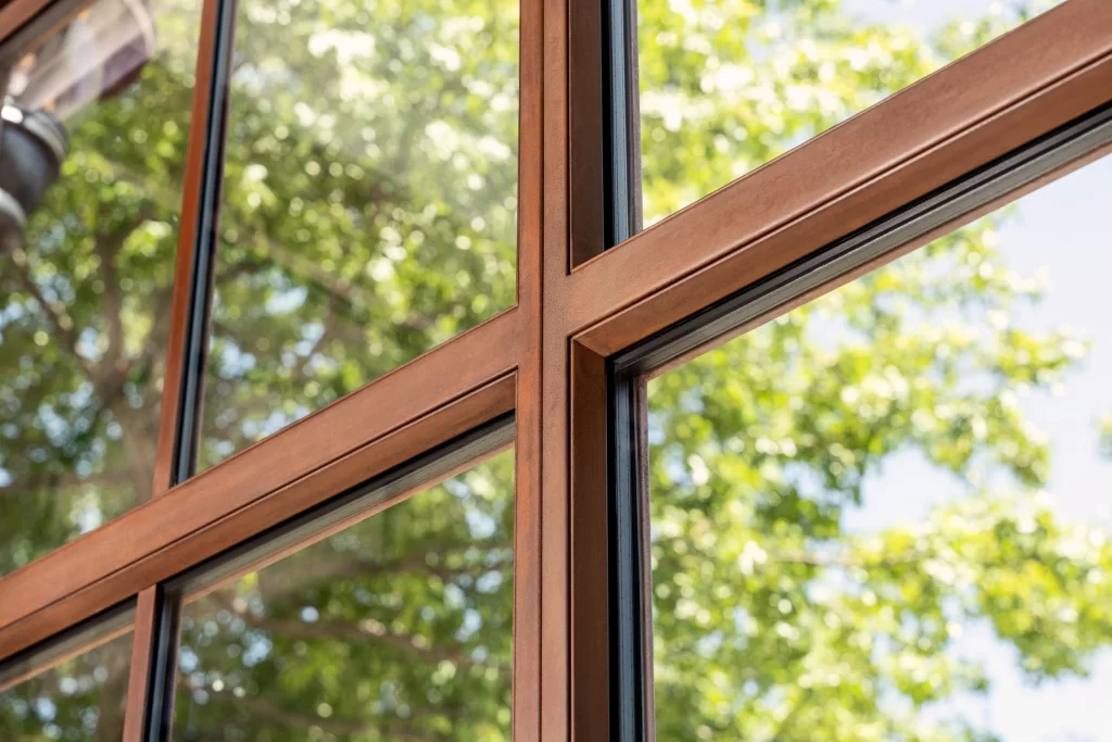 Close-up of a window corner, showcasing the warm, rich tones of the expertly crafted corten steel frame, which is perfectly polished to highlight its fine grain and joins, with clear glass panels reflecting a vibrant outdoor scene of lush green trees bathed in natural sunlight.