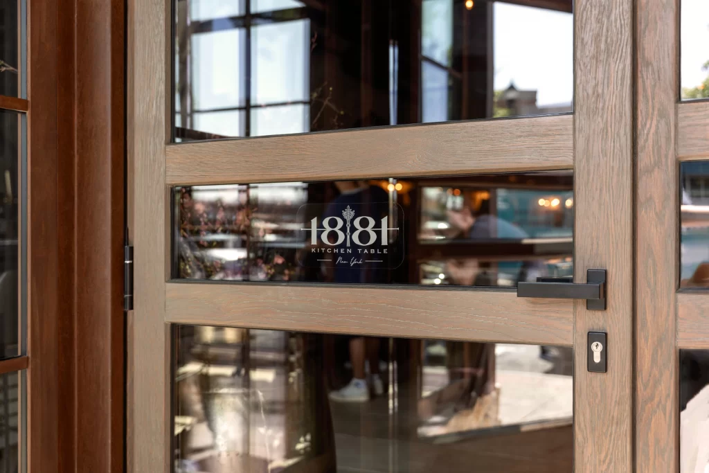 Close-up view of a beautifully crafted wooden door with a modern, light-stained finish, featuring rectangular glass inserts, a sleek black handle, and a keyhole, adorned with the elegant branding of "1818 Kitchen Table New York," reflecting the interior ambiance and exterior surroundings in the glossy glass surface.