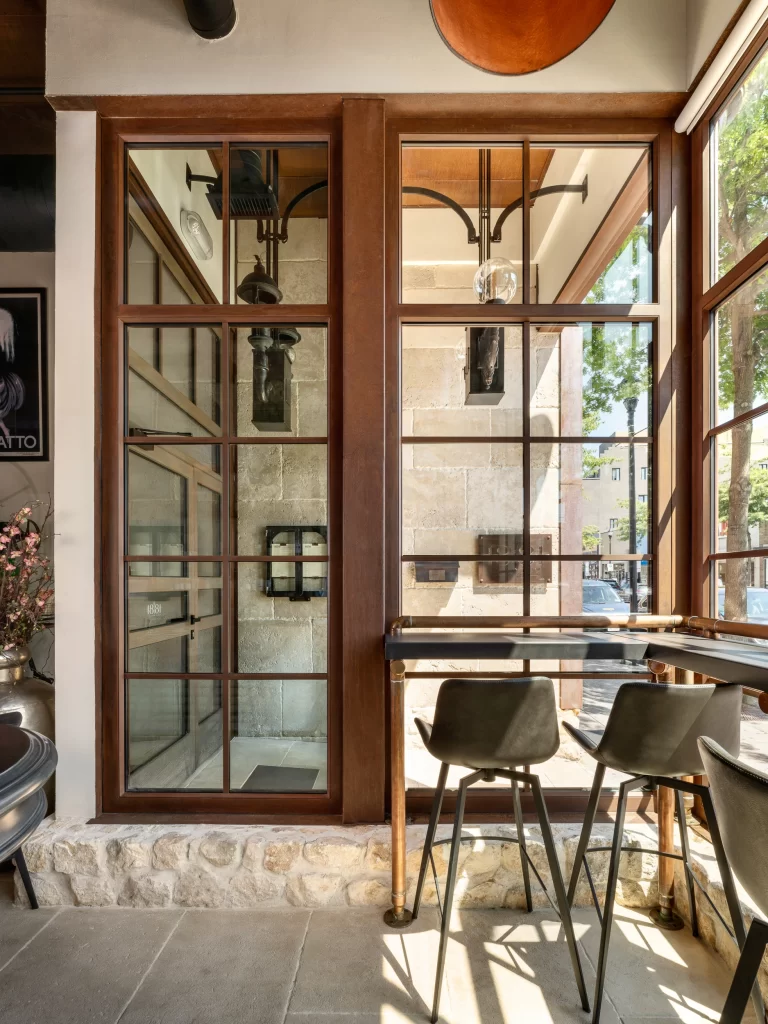 Stylish interior corner with tall, industrial-style glass and wood-framed windows that let in ample natural light, highlighting the warm tones of the stone wall and floor; a modern bar table with sleek black chairs is placed against a backdrop of polished brass fixtures, blending rustic and contemporary design elements harmoniously.