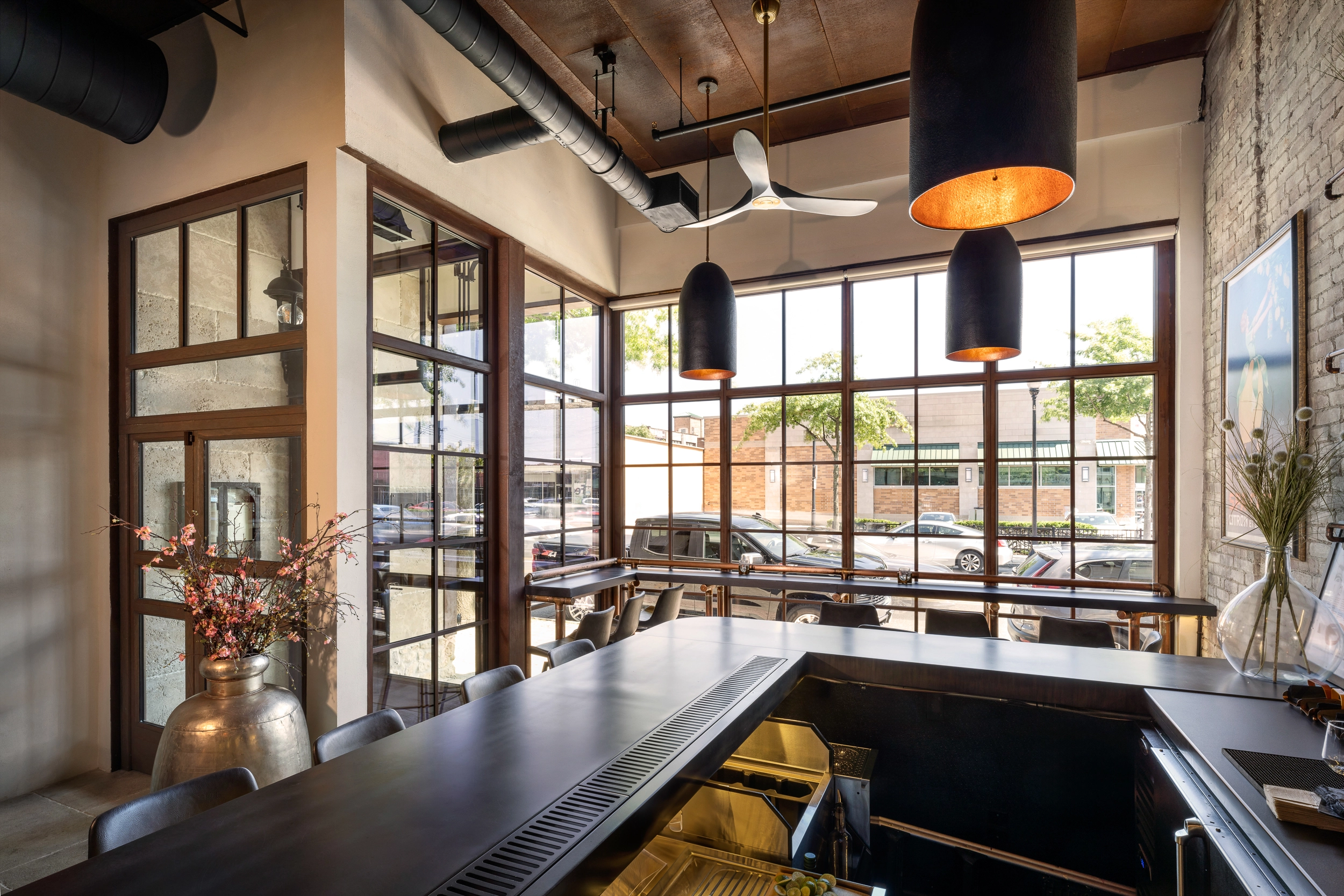 A stylish interior of a modern restaurant with large industrial-style corten steel windows providing ample natural light, black pendant lighting hanging from a wood-paneled ceiling, an exposed duct system, a sleek bar counter in the foreground, and a decorative vase with pink flowers adding a touch of elegance, while outside, a tree-lined street with parked cars and shops can be seen.