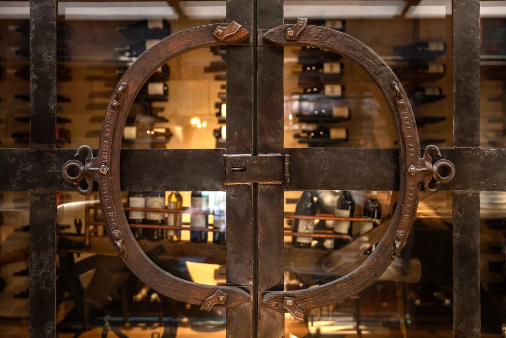 This image features a close-up view of a rustic metal wine cabinet door with an industrial-style circular iron handle, accentuated by rivet details and textured wood inlays, while the background reveals an organized wine cellar, illuminated warmly to highlight the artisanal craftsmanship of the furniture.