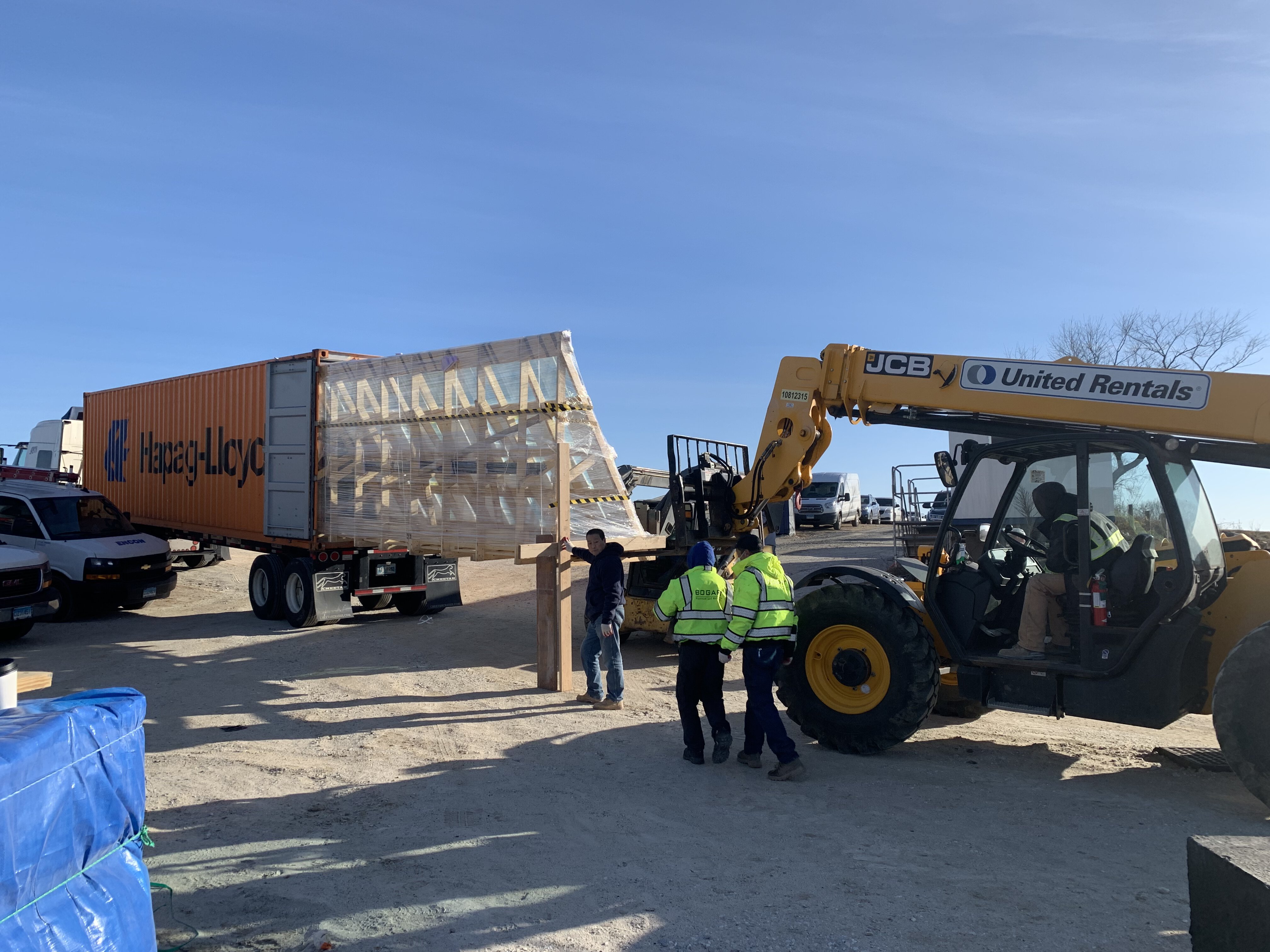 A construction site where workers are unloading a large, crated glass panel from an orange Hapag-Lloyd shipping container, a JCB telehandler, rented from United Rentals, is being used to carefully lift and transport the crate.