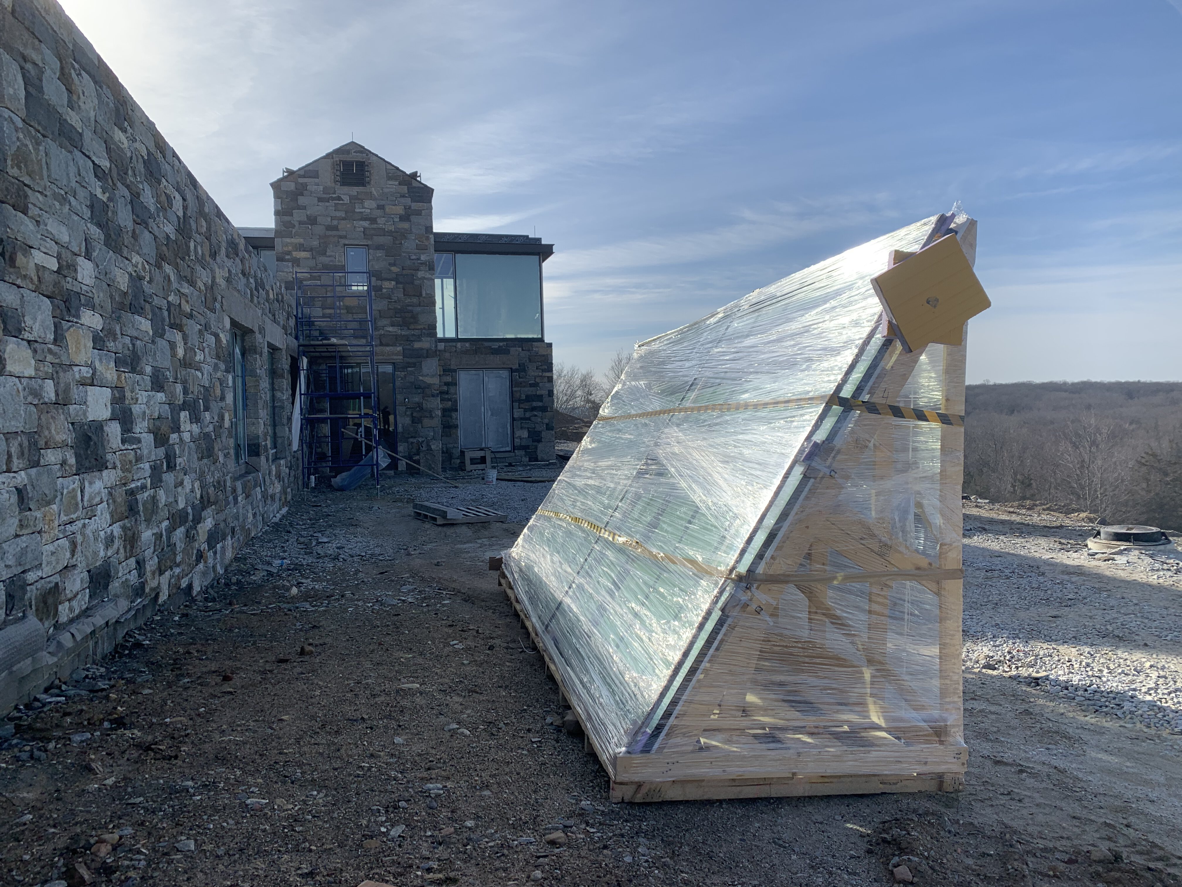 A construction site featuring a modern stone-clad building with large glass windowsand in the foreground, a large crate wrapped in plastic contains multiple glass panels, securely packed in a wooden A-frame.