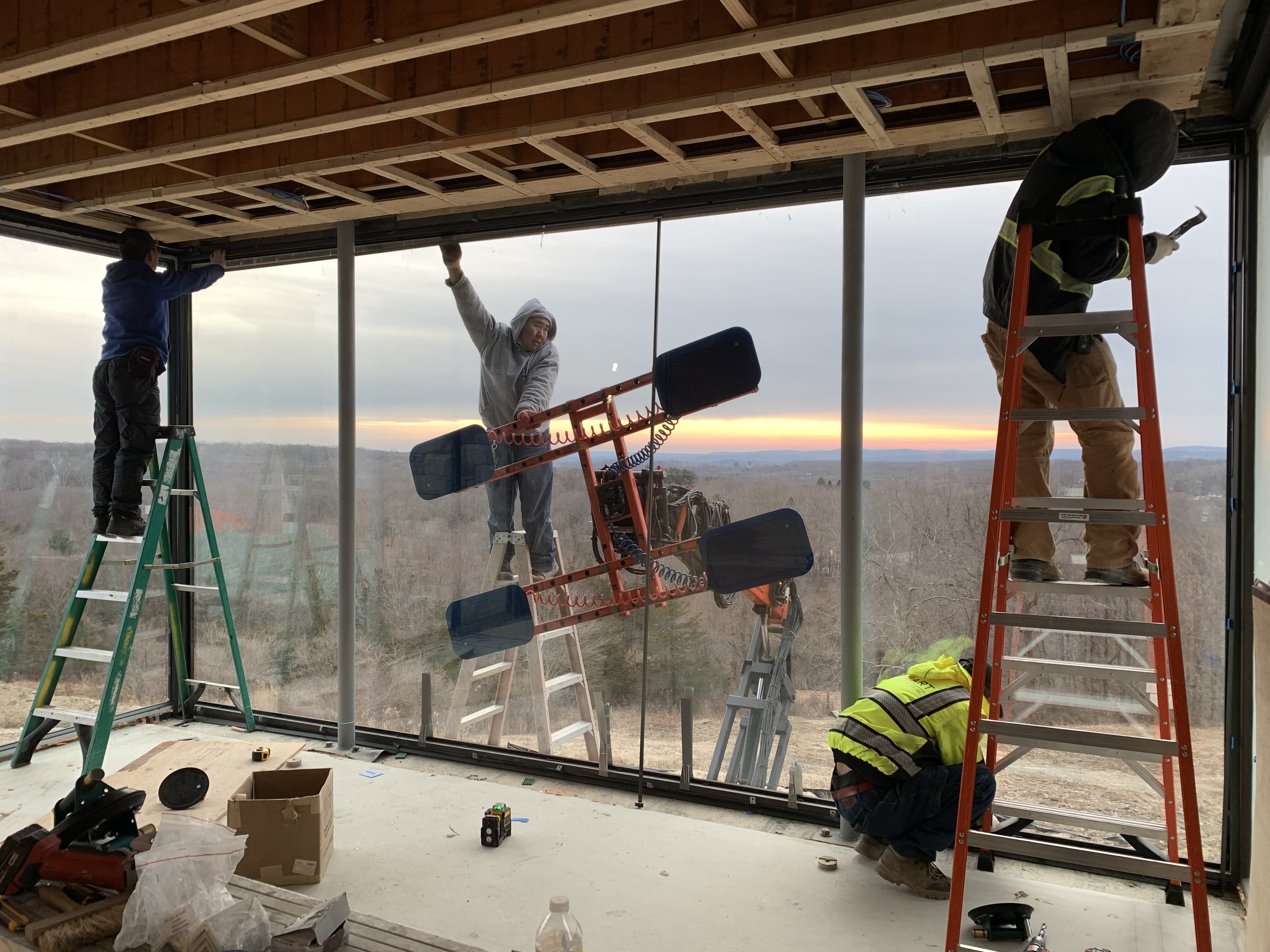 Construction workers installing a large floor-to-ceiling glass window in a modern building under construction, two workers stand on ladders inside, securing the top frame, while another worker in a safety vest kneels near the bottom track ad outside, a worker on a ladder assists with positioning the glass, supported by a suction lifter attached to heavy machinery.