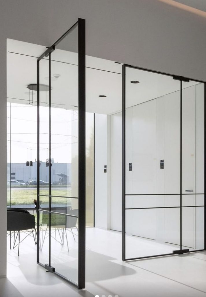 Minimalist interior space with floor-to-ceiling black-framed glass pivot doors, seamlessly integrating different areas while maintaining an open and airy feel, with sleek white walls and flooring enhancing the modern, sophisticated aesthetic, the transparent glass panels allow ample natural light to flood the space, creating a bright and inviting atmosphere, while the thin black metal frames add a striking contrast and a sense of industrial elegance