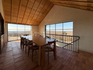 A dining space with a high vaulted wooden ceiling and large floor-to-ceiling windows that provide a panoramic view of the surrounding landscape, a black metal railing lines the edge of the space, overlooking a lower level and the natural light enhances the warm tones of the wood and earthy materials, creating a rustic yet modern aesthetic.