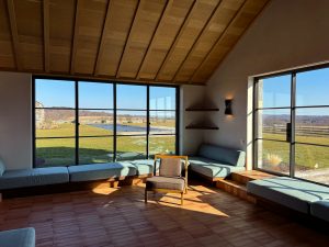 A sunlit lounge area with a vaulted wooden ceiling and terracotta tile flooring features built-in cushioned bench seating along the perimeter witht the Large floor-to-ceiling windows and glass doors provide expansive views of the surrounding landscape, including a lawn and a covered pool