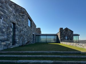 A modern stone-clad residence with a landscaped courtyard and tiered stone steps leading to a grass-covered terrace, the architecture features large glass windows and doors framed by textured stone walls, blending contemporary and rustic design elements