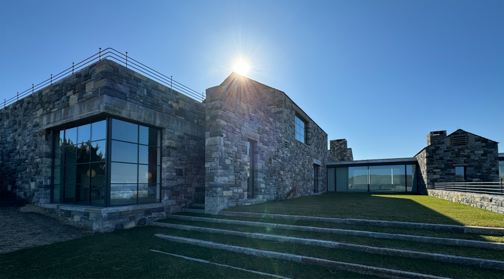 Modern stone-clad building with large glass windows, surrounded by a well-manicured lawn and tiered stone steps leading up to the structure, architectural style blends contemporary elements with rustic stone construction, featuring clean lines, metal railings, and expansive glass sections that reflect the surrounding environment.