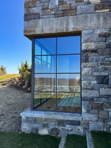 A close-up view of a large window set into a stone-clad wall, reflecting the clear blue sky and surrounding landscape, the black metal-framed glass window reveals part of the interior, showing a tiled floor and a hanging light fixture, the stone wall is composed of irregularly shaped, multi-toned stones, with a concrete lintel above the window.