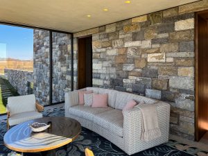 A cozy sitting area with a textured sofa adorned with soft pink pillows and a beige throw blanket, positioned against a rustic stone wall, floor-to-ceiling glass windows offer a view of the surrounding landscape, with an exterior stone-clad wall visible through the glass, recessed ceiling lights add warmth to the space, blending natural materials with modern design elements.