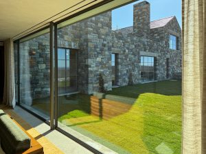 A view of a modern stone-clad residence through a floor-to-ceiling glass window, reflecting sunlight onto the green lawn outside, the building features large windows, a sloped roof, and a prominent stone chimney, sheer curtains frame the glass, and the interior's wooden furniture and warm tones contrast with the cool gray stone.