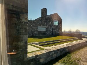 A view of a modern stone-clad residence through a floor-to-ceiling glass window, reflecting sunlight onto the green lawn outside, the building features large windows, a sloped roof, and a prominent stone chimney, sheer curtains frame the glass, and the interior's wooden furniture and warm tones contrast with the cool gray stone.