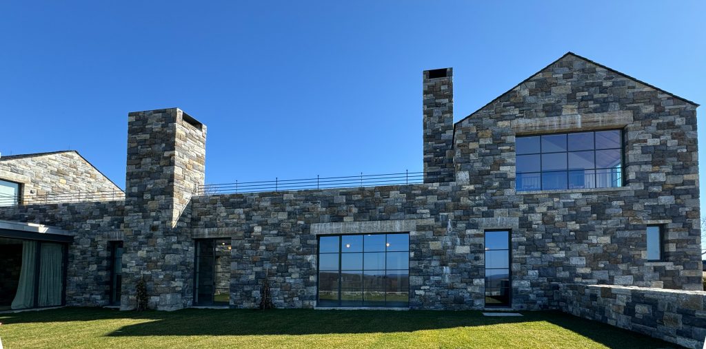 Modern stone-clad house with a rustic yet contemporary aesthetic, featuring a facade composed of irregularly shaped, multi-toned gray stone blocks that create a natural and textured appearance, complemented by large black-framed windows and doors that allow ample natural light to enter, the black metal-framed glass elements seamlessly integrate with the structure, providing a modern touch that balances the traditional stonework, while exterior lighting fixtures inside the home are faintly visible through the windows, adding warmth to the overall composition.