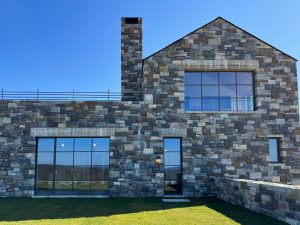 A modern stone-clad building with large black-framed windows and a tall chimney set against a clear blue sky. The facade features a mix of rectangular and square stone blocks in varying shades of gray and beige, with the glass door and two large windows reflect the surrounding landscape, while an upper-level window reveals an interior space with a vaulted wooden ceiling, a metal railing runs along the rooftop, and the neatly trimmed grass in the foreground contrasts with the rugged stone exterior.