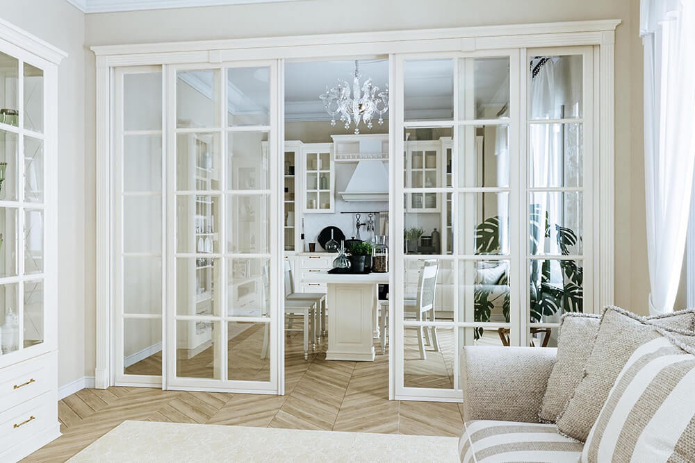 An elegant, light-filled interior featuring a set of white-framed French sliding doors with multiple glass panels, which separate the living room from the kitchen and dining area while maintaining an open and airy feel