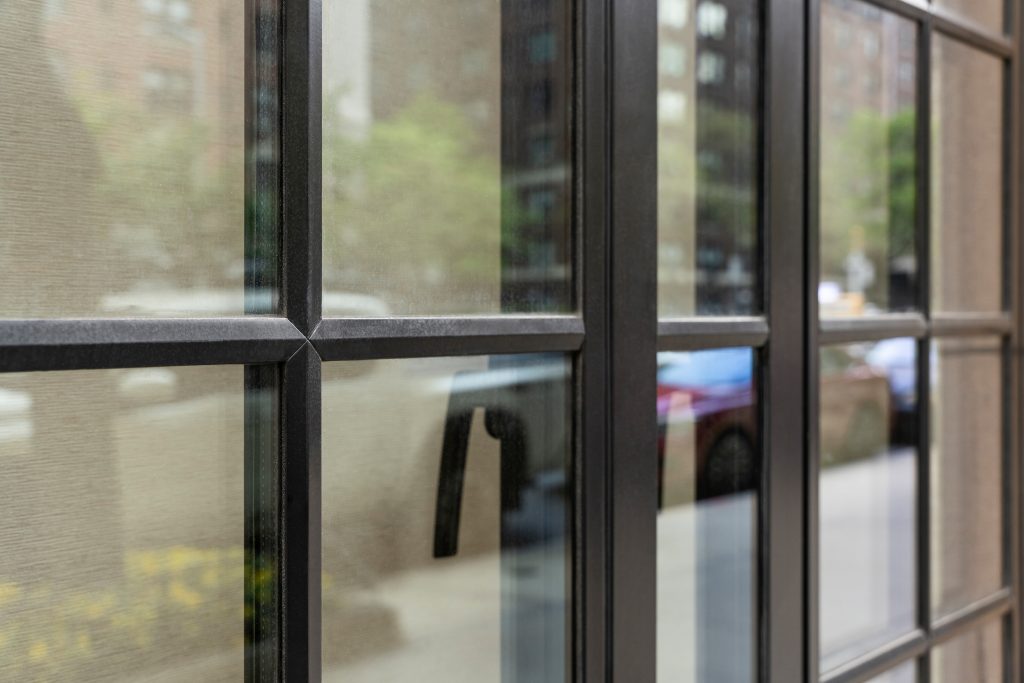 Close-up of a black-framed glass door with a grid pattern, showcasing the intersection of the metal framework and the clean, reflective glass panels, which offer a glimpse of the urban environment outside, the textured metal finish gives a sophisticated industrial touch, while the glass reflects the surrounding buildings, greenery, and parked cars, creating a dynamic interplay of interior and exterior elements