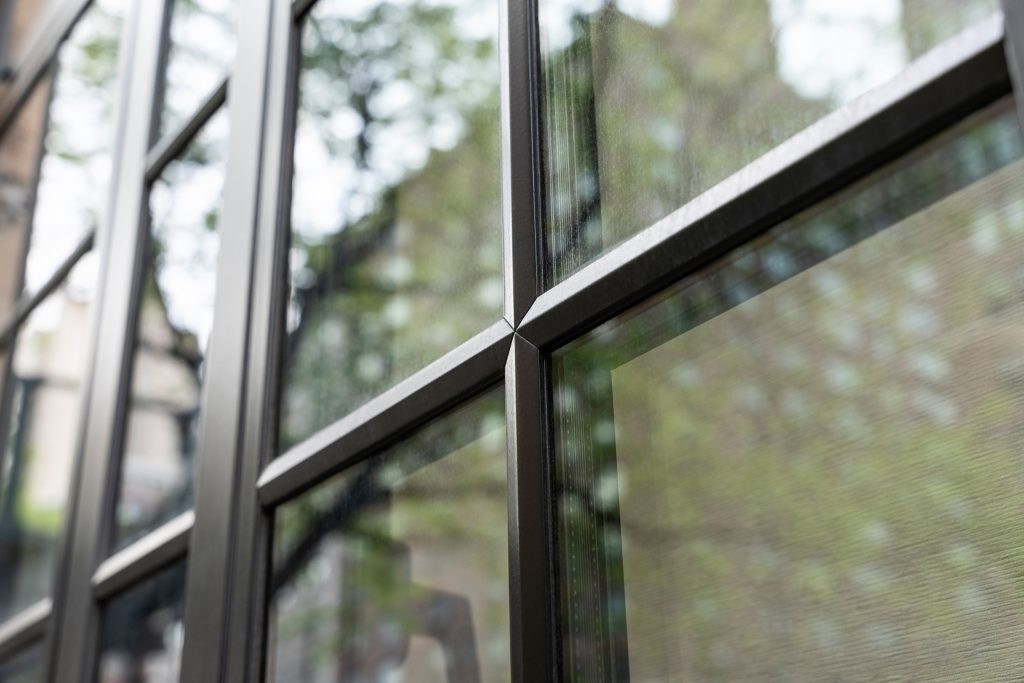 Close-up of a black-framed glass window or door, highlighting the precise metal grid framework and the seamless intersection of the glass panes, which reflect the surrounding urban environment, including trees, buildings, and soft natural light