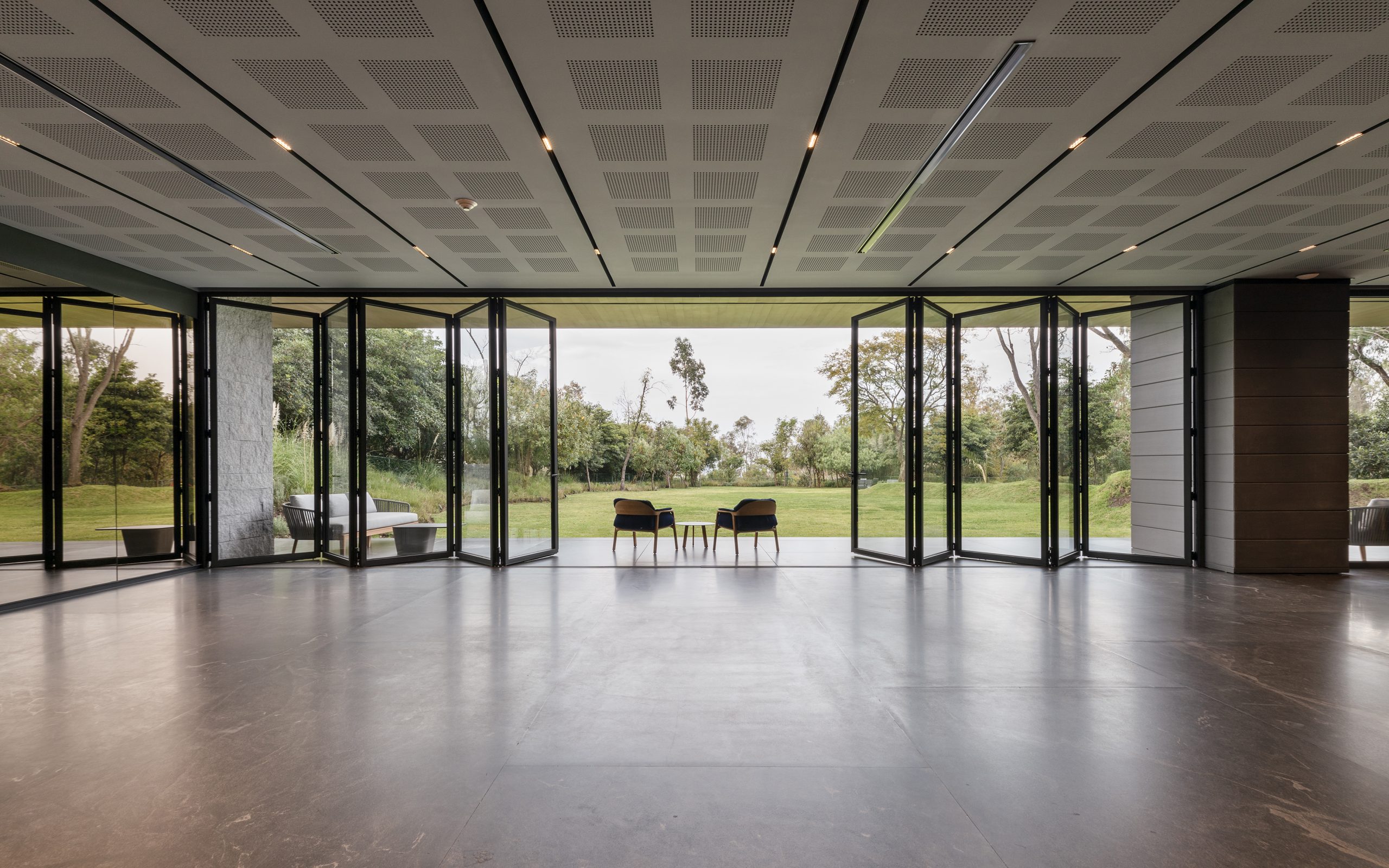 A spacious modern interior with a dark polished floor and a ceiling adorned with perforated acoustic panels is seamlessly connected to the outdoors through large black-framed bi-fold glass doors, which are fully opened to reveal a lush green landscape with scattered trees, a manicured lawn, and a cloudy sky, while two chairs with a small round table between them are symmetrically placed at the center of the opening, offering a serene and inviting transition between the sleek indoor space and the natural beauty beyond, complemented by additional outdoor seating arrangements on either side, partially visible through the expansive glass walls