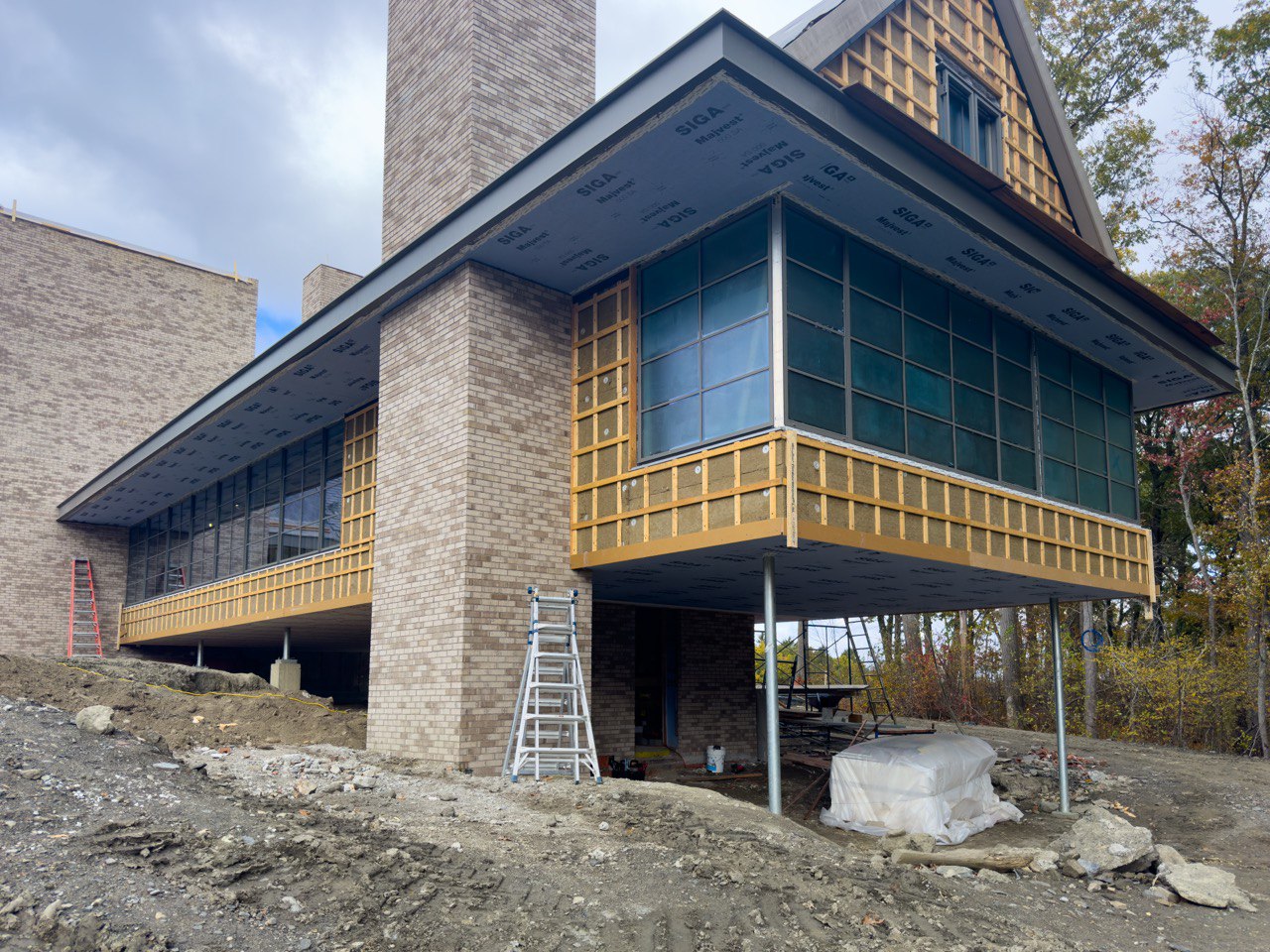 A modern multi-level building under construction features a combination of brick and glass elements, with sections of its exterior still being completed, evident from the exposed wooden furring strips and protective SIGA membrane covering parts of the walls and soffits, while large glass windows span across the structure, some of which appear to be covered with temporary protective film, and an overhanging section of the building is supported by steel columns, creating a floating effect, as the surrounding area consists of an uneven dirt surface with construction materials, ladders, scaffolding, and tools scattered around, indicating an active worksite, all set against a backdrop of trees with autumn foliage under a cloudy sky