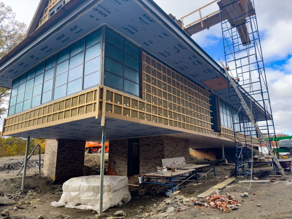 A modern building under construction features an elevated cantilevered section supported by steel columns, with a combination of brick and large steel windows & doors, while exposed wooden furring strips and SIGA membrane indicate ongoing exterior finishing work, and a tall scaffolding structure extends to the roof, where temporary wooden planks suggest additional construction activities, as the ground below is strewn with bricks, construction materials, and equipment, including a wrapped pallet, workbenches, and a partially visible orange construction vehicle, all set against a backdrop of autumn trees and a partly cloudy sky.