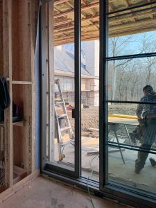 A partially constructed interior space with exposed wooden framing, insulation, and electrical wiring is visible, featuring a large sliding glass door that provides a view of an outdoor construction area, where a worker in a plaid shirt and jeans is engaged in a task and in the background, a modern brick house with large windows and scaffolding surrounding it stands among leafless trees under a clear blue sky.