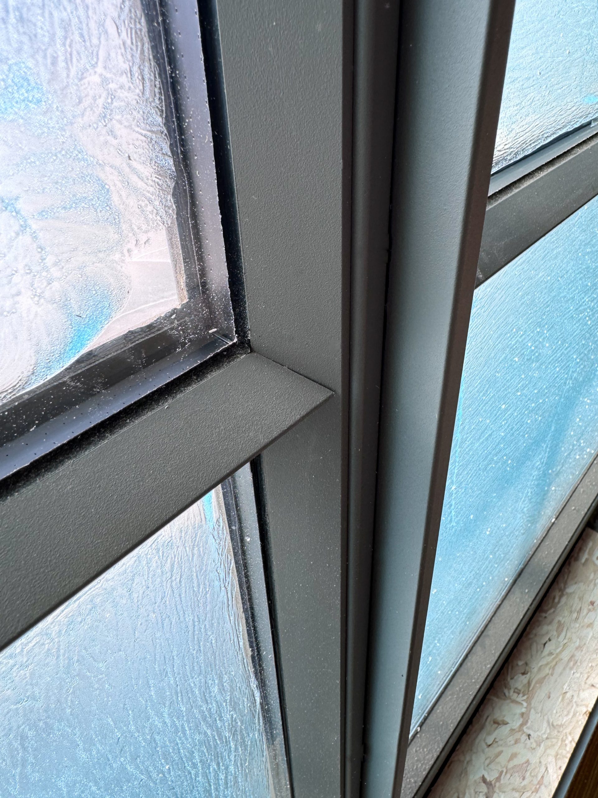 A close-up view of a modern steel-framed window corner highlights the precise intersection of the dark metal frame, which features a matte finish and sharp edges, with the textured, semi-opaque glass panes that have a frosted appearance with condensation or protective film residue, while small dust particles and construction debris are visible on the frame, suggesting recent installation or ongoing construction