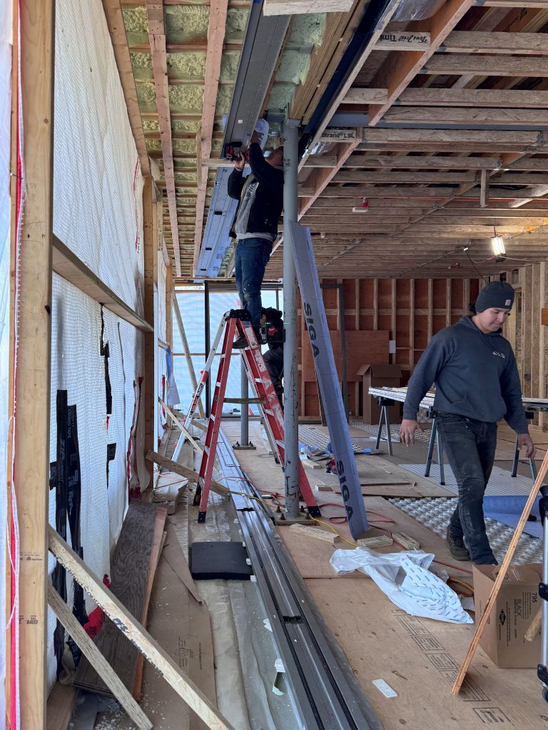 A construction site with an unfinished interior steel structure is shown, where two workers are engaged in tasks, one standing on a red ladder while using a power drill to secure a metal beam to the ceiling framework, and another in a dark hoodie and beanie walking across the plywood-covered floor, surrounded by tools, and construction materials.