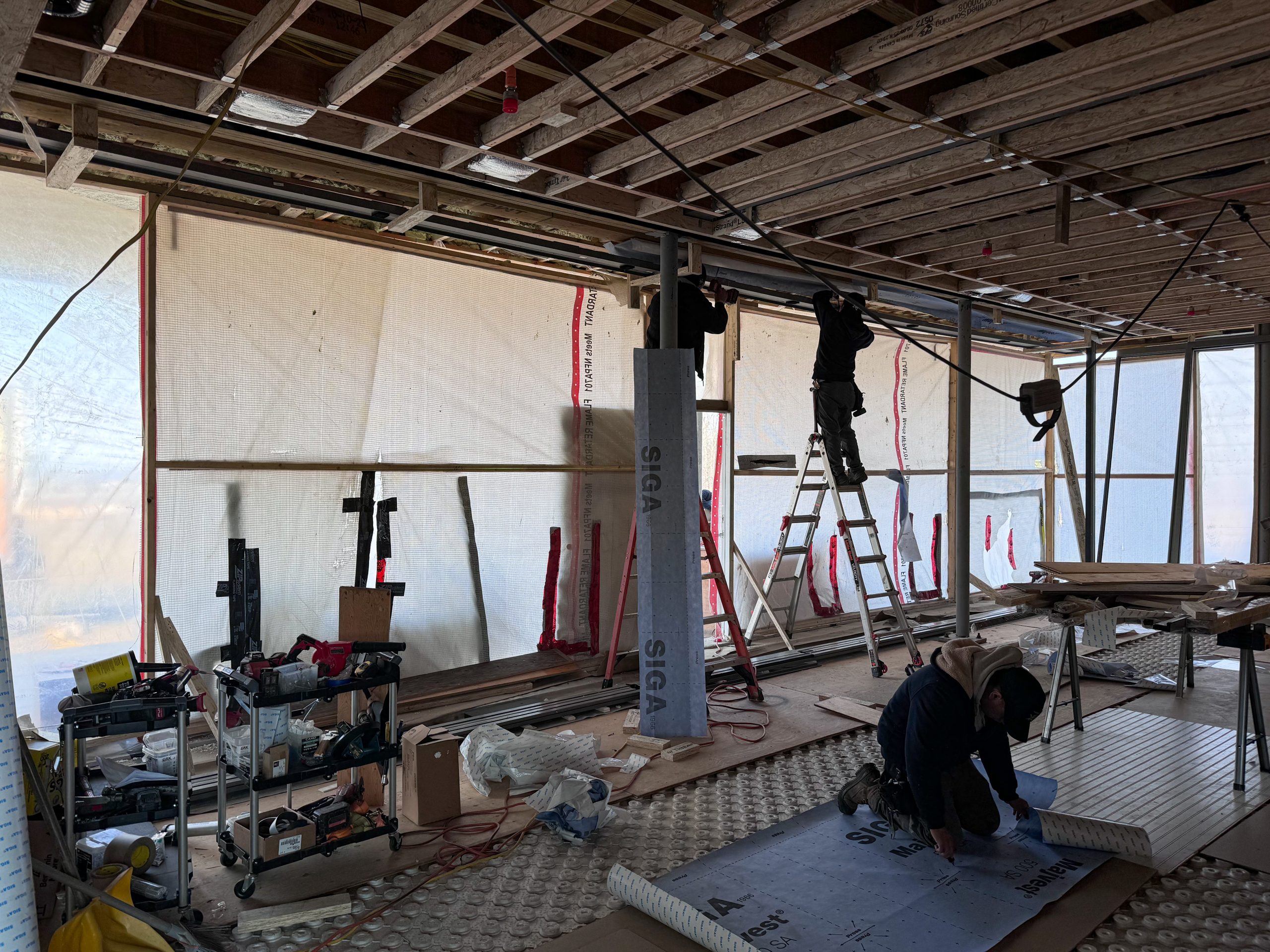 An active construction site inside a building features exposed wooden ceiling beams with electrical wiring running across, a worker standing on a ladder installing or adjusting components near the ceiling, another worker kneeling on the floor securing a sheet of insulation material while large translucent plastic sheets cover the walls, allowing diffused natural light to enter the space, creating an industrial atmosphere with a mix of organization and ongoing work.