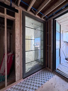 In an unfinished interior space is black-framed window with horizontal mullions and protective plastic film offers a view of the outside, next to a partially open dark grey pivot door glass door that leads to an exterior space.