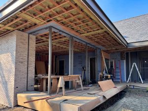 A partially constructed modern residential building features an exposed wooden ceiling grid with spray foam insulation between the beams, supported by steel columns, into which steel windows & doors will be installed later, extending from a partially completed concrete patio, while the exterior walls combine light-colored brick and weather-resistant barrier wrap branded and various construction materials, including wooden planks, sawhorses, and tools, are scattered throughout the site, with a worker wearing a dark hoodie and pants seen near a red ladder, actively engaged in the construction process.