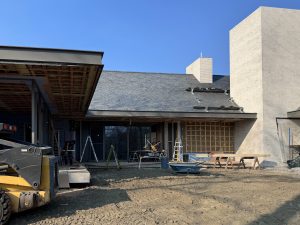 A modern residential building under construction features a large sloped roof with dark shingles, partially installed roofing materials, and a combination of light-colored brick and unfinished exterior walls covered in weather-resistant sheathing, with an extended covered patio supported by steel columns and an exposed wooden ceiling structure filled with insulation, while multiple ladders, sawhorses, and scattered construction tools are present, along with workers engaged in various tasks and a yellow construction vehicle partially visible in the foreground on a dirt-covered ground