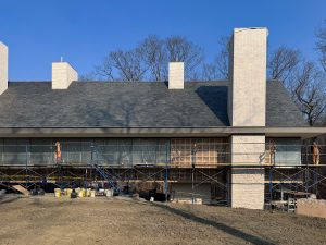 A large, modern house under construction features a steeply pitched dark gray shingle roof, multiple tall and rectangular white brick chimneys, and a facade partially covered in scaffolding with protective plastic sheets over the windows, with workers present on the scaffolding performing tasks, and a backdrop of leafless trees under a clear blue sky indicating a winter or early spring setting.