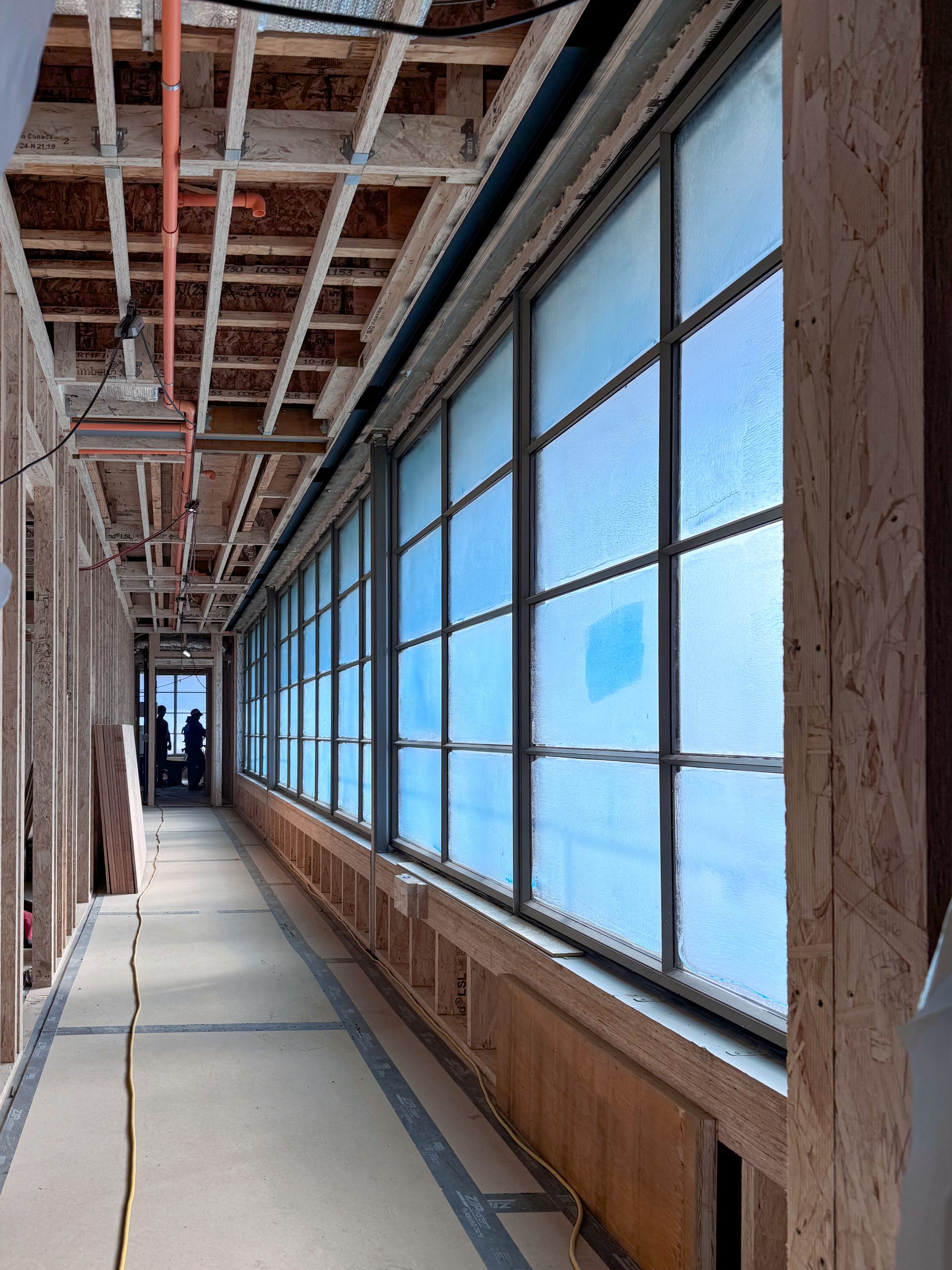 A long, narrow hallway under construction features an unfinished ceiling with exposed wooden beams, metal brackets, and electrical wiring, while the right side is lined with large industrial-style steel windows covered with semi-transparent blue protective film, framed by dark grey steel mullions, and the lower portion of the wall is partially enclosed with wooden panels.