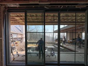 A large glass sliding door with dark grey metal frames reflects the partially constructed interior of a modern house, where exposed wooden ceiling beams and insulation are visible, while beyond the glass, a construction site extends to an outdoor area where multiple workers are engaged in various tasks, with construction materials, ladders, and wooden planks surrounding the workspace, and a background featuring a leafless forest and another section of the house with scaffolding, suggesting an ongoing residential project