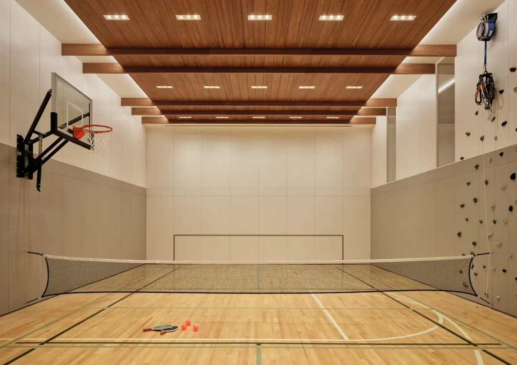 This indoor court is next-level impressive—a sleek, multi-purpose sports space with a polished wood floor featuring court markings for basketball and pickleball, it’s equipped with a basketball hoop, a net set up across the center, and even a rock climbing wall with an auto-belay system on the side while the ceiling’s wood paneling and exposed beams add a warm, upscale touch, while built-in lighting keeps it bright and functional. 
