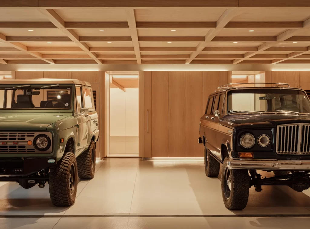 Stunning darage Iooks like a luxury showroom than a traditional garage—perfectly lit with soft, recessed lighting and a coffered wood ceiling that adds elegance, the space features two vintage SUVs, a classic green Ford Bronco and a dark Jeep Wagoneer, both in pristine condition.