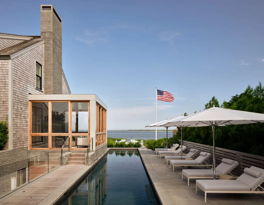 A serene coastal residence features a narrow rectangular infinity pool bordered by a wooden deck walkway and a row of plush white cushioned lounge chairs shaded by large white umbrellas, with a tall American flag fluttering in the breeze against a clear blue sky and the tranquil ocean visible in the background, while the main house clad in weathered cedar shingles includes a modern glass-walled sunroom with natural wood framing that opens onto the pool area via a small stairway with glass railings, surrounded by manicured greenery and coastal landscape elements.